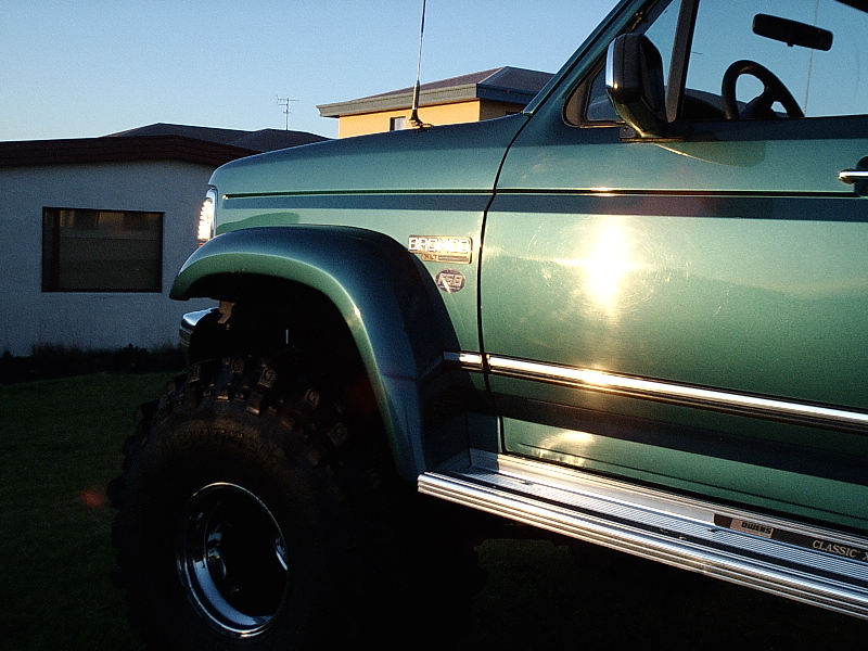 Green lifted Ford Bronco front fender with large mud tire, low-angle side view at sunset by a house