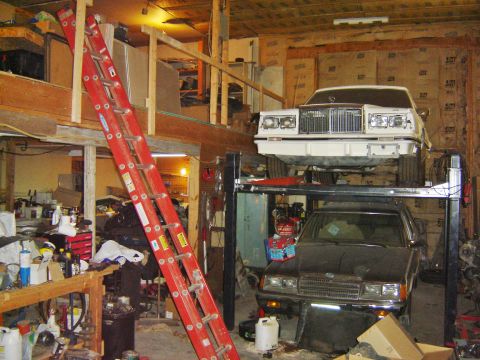1984 Ford Mustang - A white sedan is elevated on a car lift in a cluttered garage, with another vehicle below.