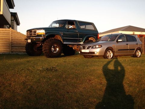 1996 Ford Bronco - A heavily modified green SUV with large tires sits beside a silver hatchback in front of houses.