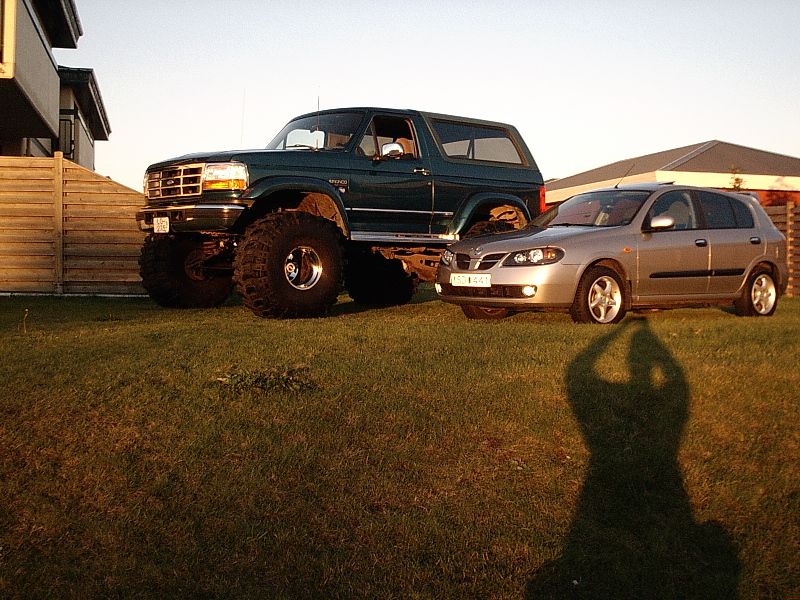 Lifted dark green Ford Bronco on big tires next to a silver hatchback on grass at dusk.