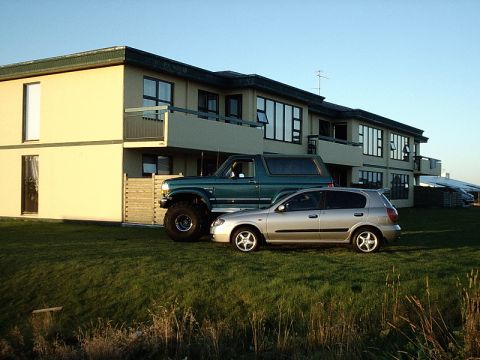 1996 Ford Bronco - A modified green Ford Bronco and a silver hatchback sit on a grassy lawn in front of a modern building.