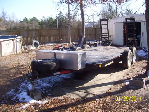 1978 Ford Bronco - Tandem-axle flatbed car-hauler trailer parked on a snow-dusted gravel and concrete pad, shot three-quarter from the front-left. It has a wo…