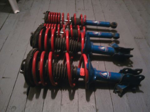 1978 Ford Bronco - Set of three blue coilover shock absorbers with red springs, possibly for a performance vehicle. One unit has an orange tag attached.