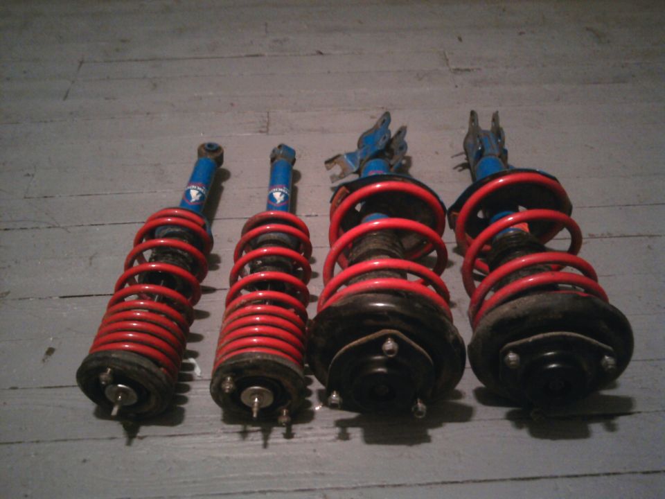 Four shock absorbers with blue bodies and red coil springs laid out on a wooden floor