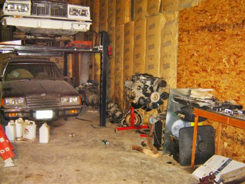 1984 Ford Mustang - Home workshop scene with a maroon sedan parked at left, a light-colored vehicle raised on a two-post lift, and a V8 engine mounted on a red…