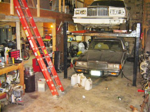 1984 Ford Mustang - Two cars are stored on a car lift in a cluttered garage with a red ladder nearby.