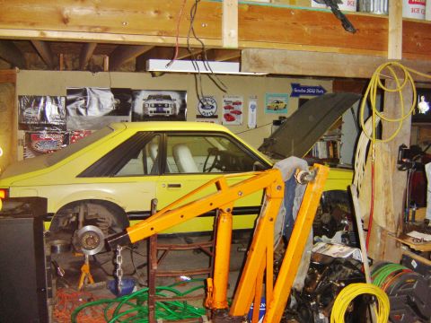 1984 Ford Mustang - A yellow Fox-body Mustang notchback sits mid-teardown in a wood-framed shop, hood raised and front wheels removed with a bare brake rotor a…