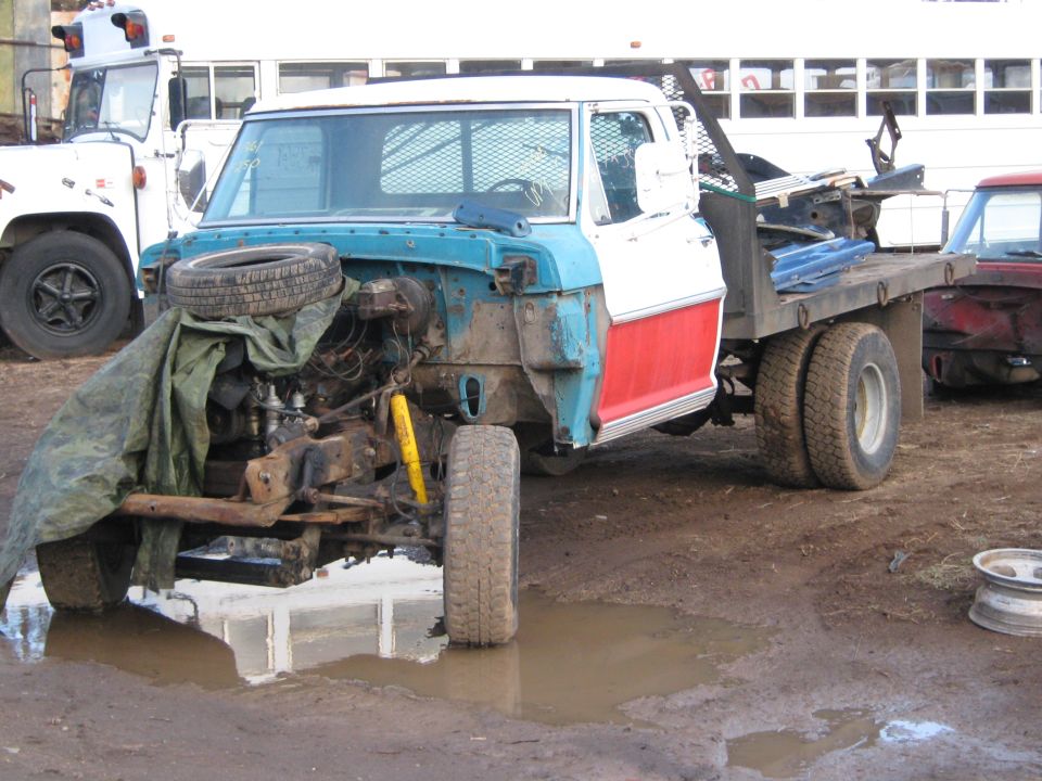 Flatbed truck missing its front clip, engine exposed, parked in mud next to another truck