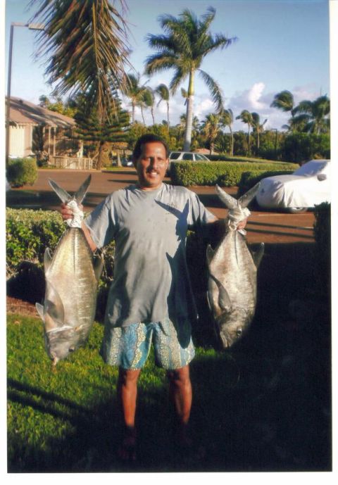 1991 Ford Bronco - A barefoot man in a gray tee and blue board shorts holds up two large silver ulua (giant trevally) on a lawn in a palm-lined tropical neigh…