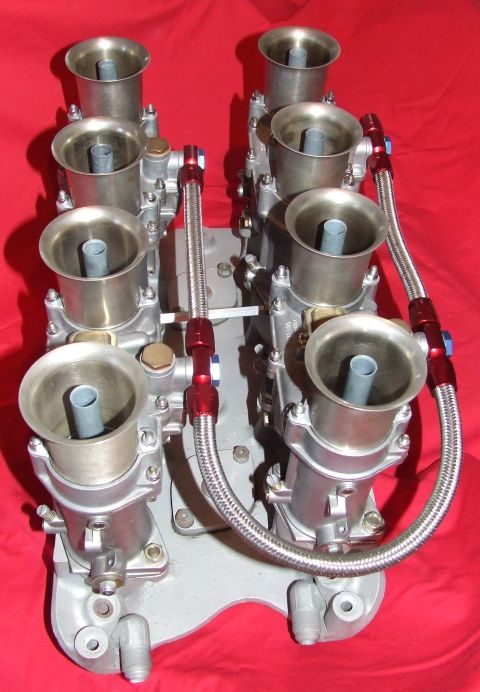 1973 Ford Mustang - Close-up of a dual quad carburetor setup with braided fuel lines against a red background.