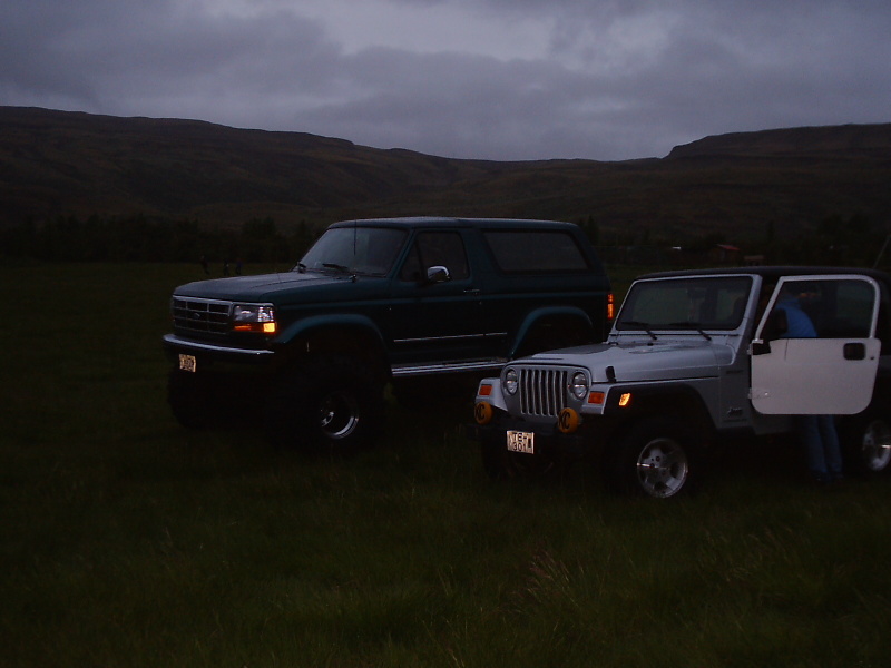 Green lifted Ford Bronco parked next to a silver Jeep Wrangler in a field at dusk