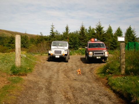 1996 Ford Bronco - Two SUVs are parked on a gravel road, separated by a chain and wooden posts.