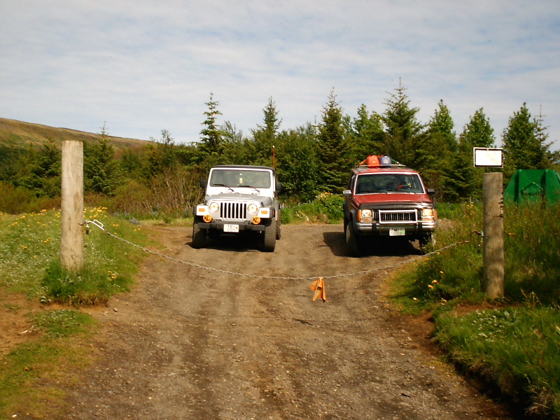 White Jeep Wrangler and red Jeep Cherokee parked on gravel behind a chain barrier