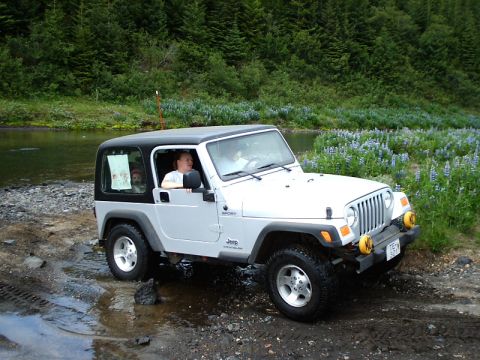 1996 Ford Bronco - A silver Jeep Wrangler navigates a muddy path beside a river, with a passenger and a child visible inside.