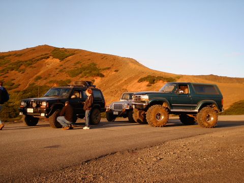 1996 Ford Bronco - Three modified SUVs, including a 1996 Ford Bronco, are parked on the side of a road with a hill in the background.