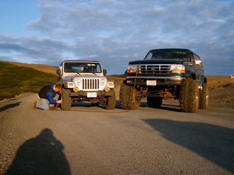 1996 Ford Bronco - A man kneels to inspect the tire of a Jeep while a large Ford Bronco sits nearby on a gravel road under a cloudy sky.