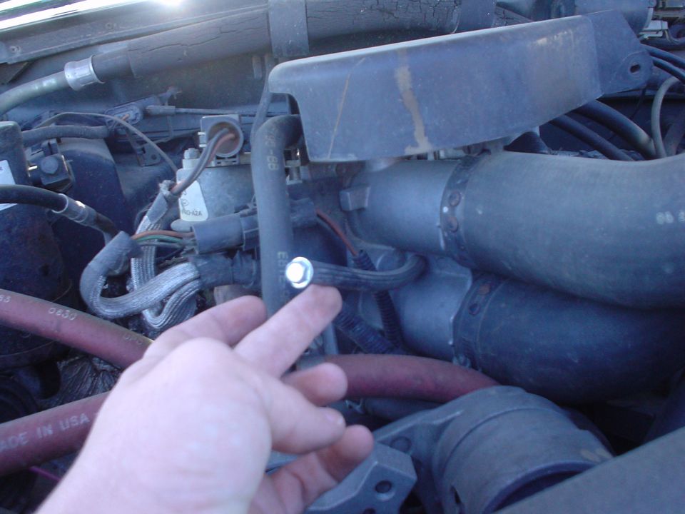 Hand pointing to a bolt among hoses and tubes in a vehicle engine bay