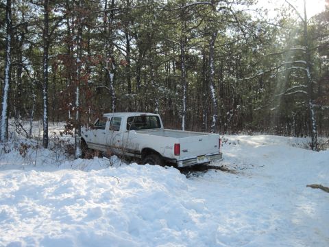 2005 Ford Excursion - A white pickup truck is stuck in a snowy wooded area, viewed from the rear.