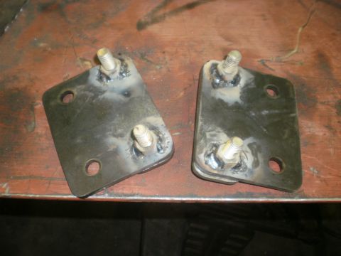 1995 Ford Bronco - A mirrored pair of fabricated steel mounting brackets sitting face-up on a rust-colored workbench, each with two threaded studs welded thro…