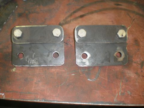 1995 Ford Bronco - A matched pair of shop-fabricated bent steel mounting brackets laid out on a rust-colored steel workbench top. Each plate has two hex bolts…