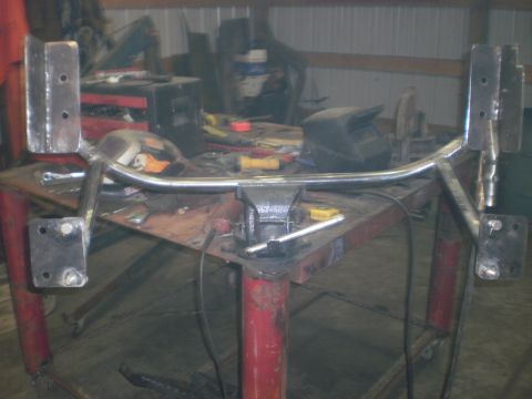 1995 Ford Bronco - A completed bare-steel tube bumper assembly with four bolt-on mounting brackets at its ends, staged on jack stands in front of a shop bench…