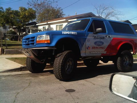 1986 Ford Bronco - A modified Ford Bronco with racing livery is seen from the front-quarter angle on a sunny day.