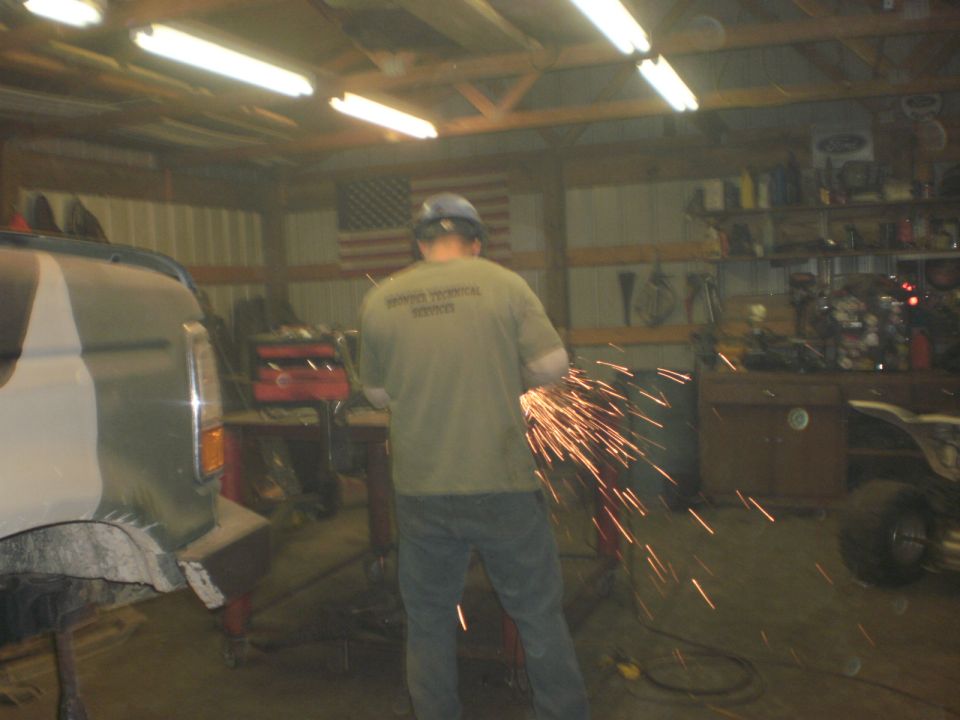 Man in a garage using an angle grinder, sparks flying. A vehicle is partially visible on the left