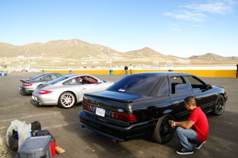 1995 Ford Taurus - A man works on a black sedan while two Porsches sit on a racetrack with mountains in the distance.