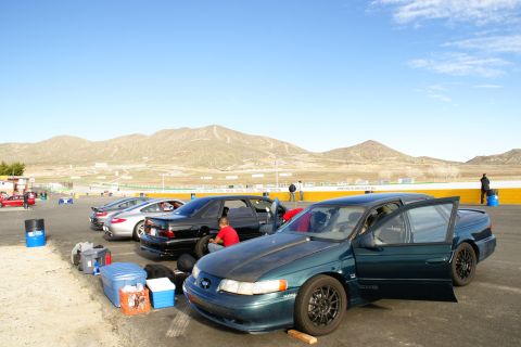 1995 Ford Taurus - A dark teal 1995 Ford Taurus sits with its doors open at a racetrack, with other vehicles and mountains visible.