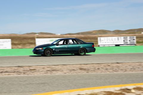 1995 Ford Taurus - A 1995 Ford Taurus sedan is captured in motion on a race track, viewed from the side under a clear blue sky.