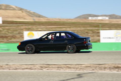 1995 Ford Taurus - Black 1995 Ford Taurus sedan seen from the side on a race track with a driver wearing a helmet.