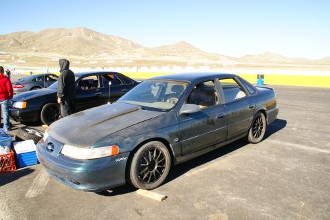 1995 Ford Taurus - A dark green 1995 Ford Taurus is parked on a race track with other vehicles and a mountainous landscape.