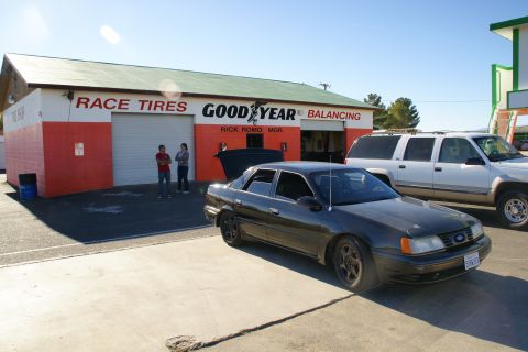 1995 Ford Taurus - A dark sedan with its trunk open is parked in front of a tire shop, with a white SUV to its right.