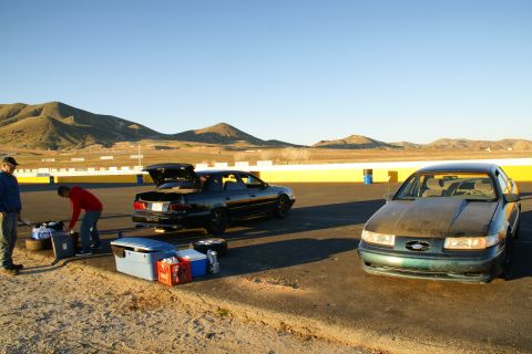 1995 Ford Taurus - Two sedans are parked on a race track with gear and people nearby.