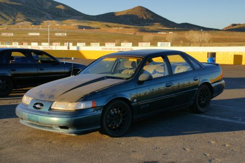 1995 Ford Taurus - Dark green 1995 Ford Taurus sedan with black wheels sits at a racetrack with another car behind it.