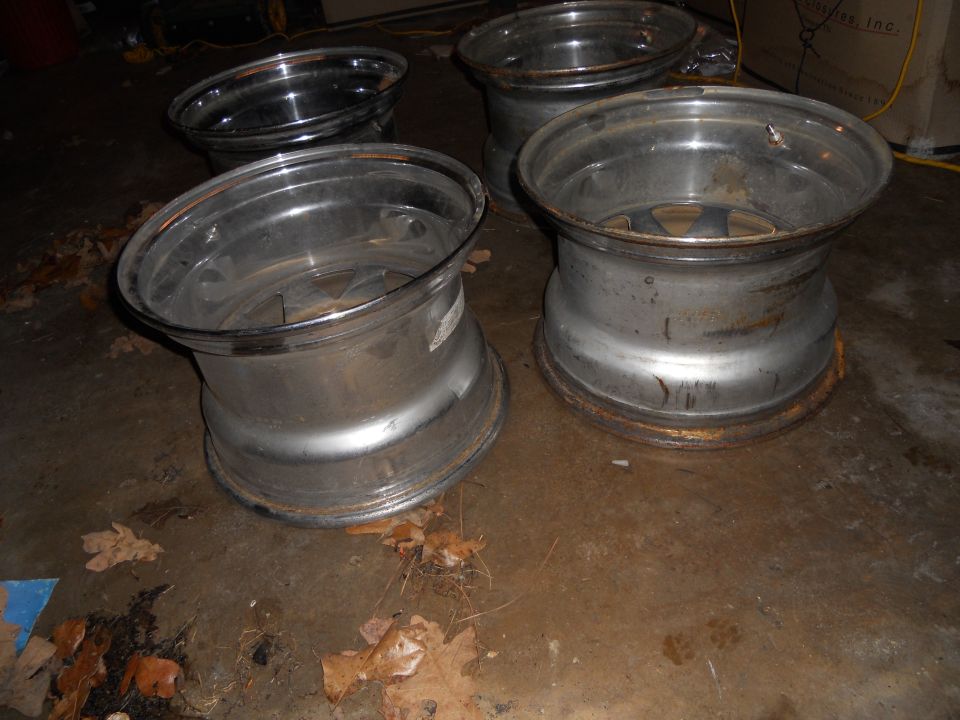 Four aluminum alloy wheels with visible rust on the rims, sitting on a concrete floor with scattered leaves