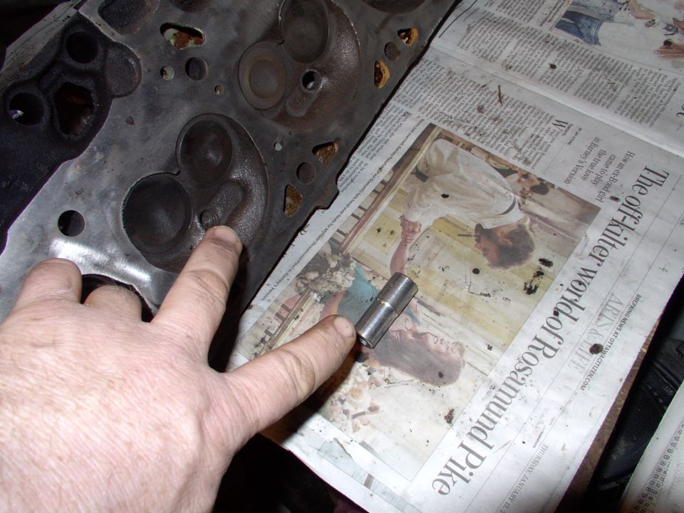 Finger pointing to combustion chamber on cylinder head over newspaper, with small steel lifter nearby