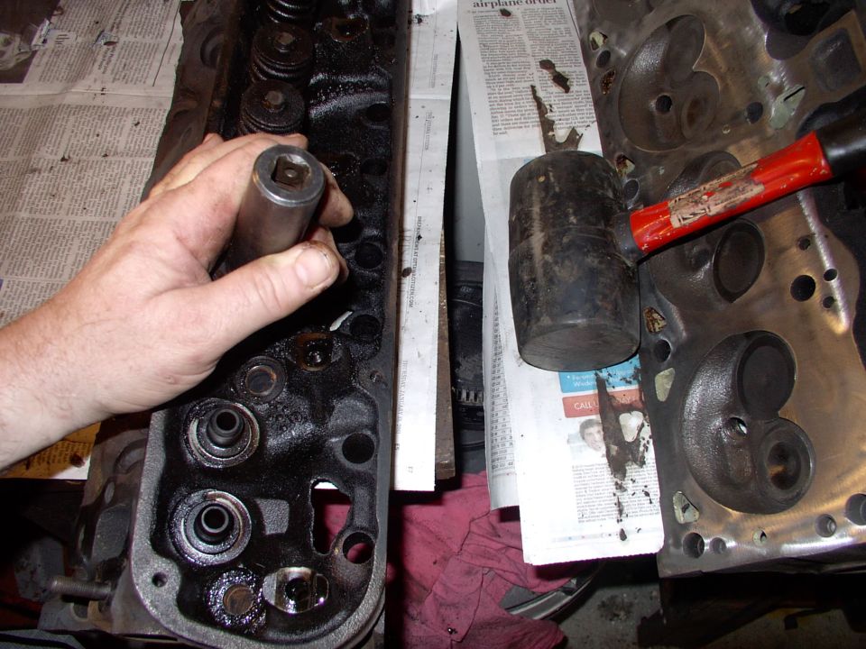 Hand holding a seal driver tool over a cylinder head valve, rubber mallet on second head nearby