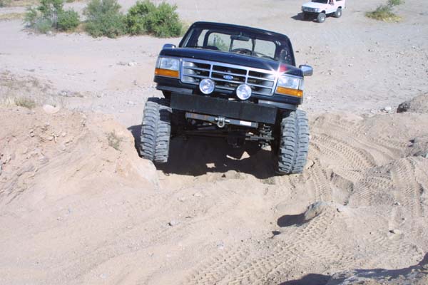 Lifted black Ford pickup climbing a dirt mound head-on, oversized mud tires flexed, auxiliary lights on the bumper
