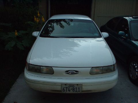 1996 Ford Taurus - A white 1996 Ford Taurus is parked in front of a garage, with another dark-colored vehicle visible to the right.
