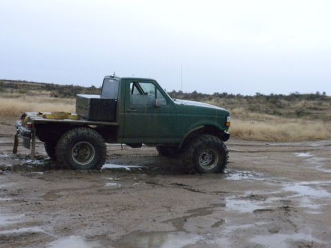 1989 Ford Bronco - Modified pickup truck with large off-road tires sits in muddy terrain under an overcast sky.