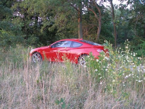 2010 Hyundai Genesis - A red 2010 Hyundai Genesis coupe is seen parked off-road in a grassy area with trees.