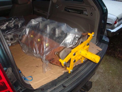 1989 Ford Bronco - Engine block in plastic wrap with a yellow hoist attachment inside a dark green SUV, with another vehicle visible to the right.