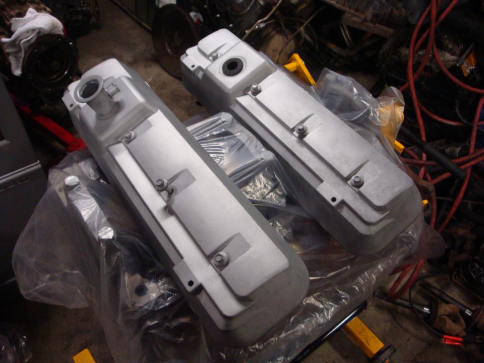 Two silver engine valve covers on a workbench, covered in plastic wrap