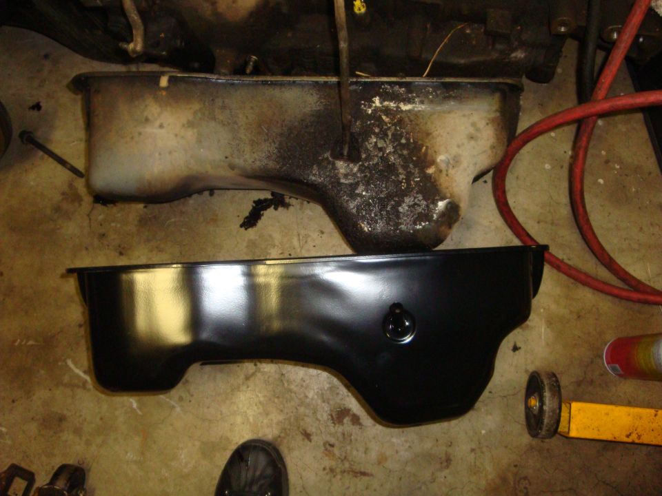 Old sludge-covered V8 oil pan next to new gloss black replacement pan on garage floor