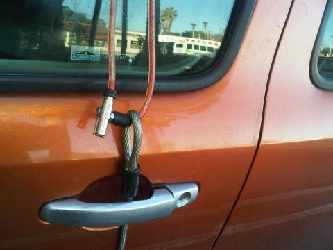 1986 Ford Bronco - Cable lock on an orange vehicle door, with reflections of buildings and a bus in the window.