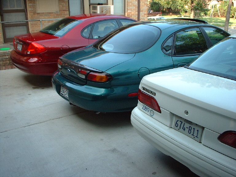 Three sedans parked in a lot. The closest is white, the middle is teal, and the furthest is red. The teal car has 'TAURUS' badging