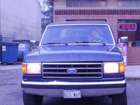 1989 Ford Bronco - This blue Ford Bronco is parked outside a building with an 'OPEN' sign, showing its front grille and headlights.
