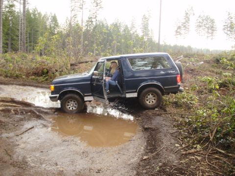 1989 Ford Bronco - A dark blue Ford Bronco navigates a muddy obstacle course in a wooded area.