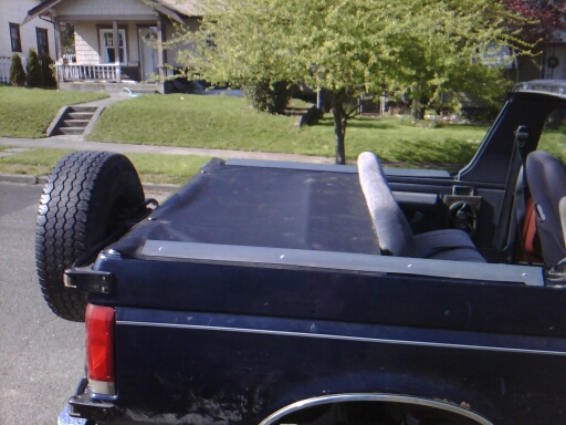 Dark blue Ford Bronco with top off, black tonneau cover over bed, rear-mounted spare tire, parked on street